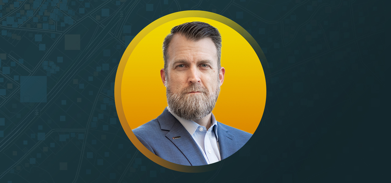 노란색과 파란색 배경에 중첩되어 비즈니스 정장을 입고 웃고 있는 Darin Welch 