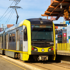 A yellow and black Metro Rail car idles at a stop on an urban street lined with skyscrapers