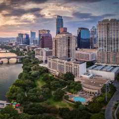 A wide-view photo of a sprawling city with a bridge over a rippling river under a pastel cloud-strewn sunset sky